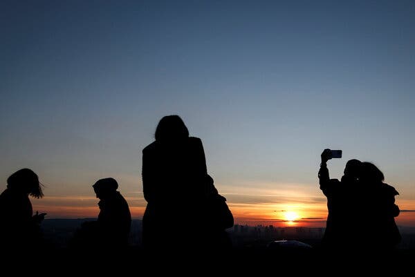 Several people, in shadow, watch a sunset. Two on the right take a selfie with a smartphone.